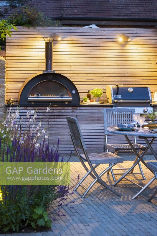 Illuminated outdoor dining area with wooden table, chairs and bespoke kitchen unit with integrated pizza oven and bbq grill at night. 