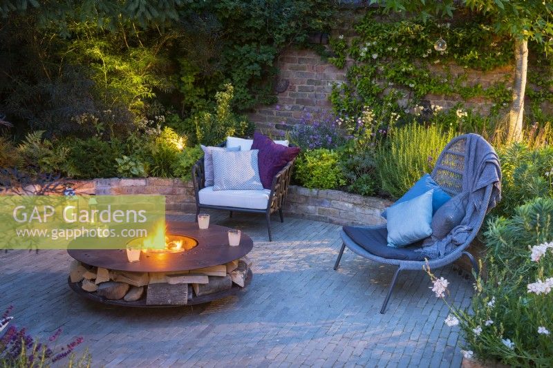 Illuminated sunken seating area with freestanding chairs and lighted fire pit at night. 