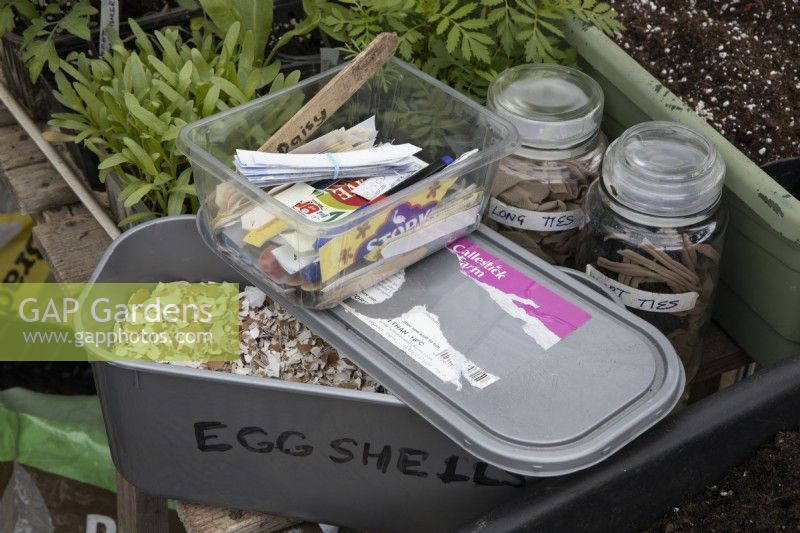 An old commercial ice cream tub is repurposed as a storage container for crushed egg shells. On top of the container sits a tub of recycled plant labels cut from old margarine tubs. Beside the container are two mason jars filled with plant ties made from cut up tights. 