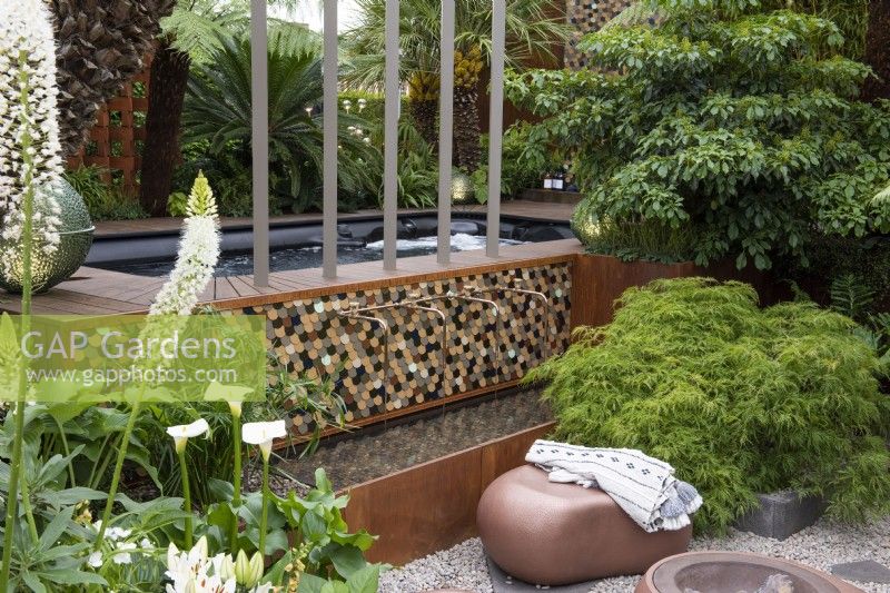A small contemporary garden incorporates a jacuzzi with a sunken area housing a fire pit with seating and a water feature. It is enclosed in maples, tree ferns, palms, a wheel tree and white flowers.