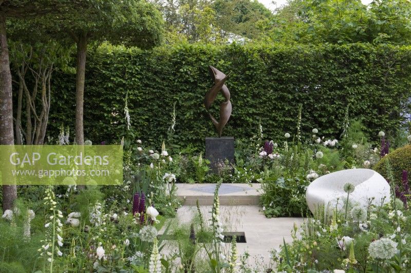 A classical, contemporary space features 'Dancers', a sculpture by Jack Eagan, offset against hornbeam hedging, amidst white and green themed borders planted with alliums, foxgloves and peonies.