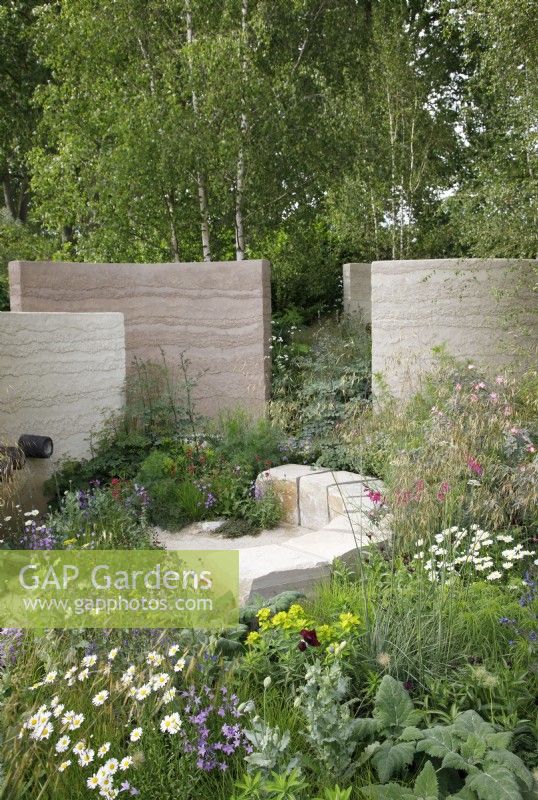 View of a seating area in The Mind Garden with large purbeck stone blocks and opposite, a water feature with ceramic water chutes encased in a clay rendered wall, the meadow planting includes Leucanthemum vulgare, Gladiolus communis subsp. byzantinus, Euphorbia palustris and Stipa gigantea - Designer: Andy Sturgeon - Sponsor: Project Giving Back.