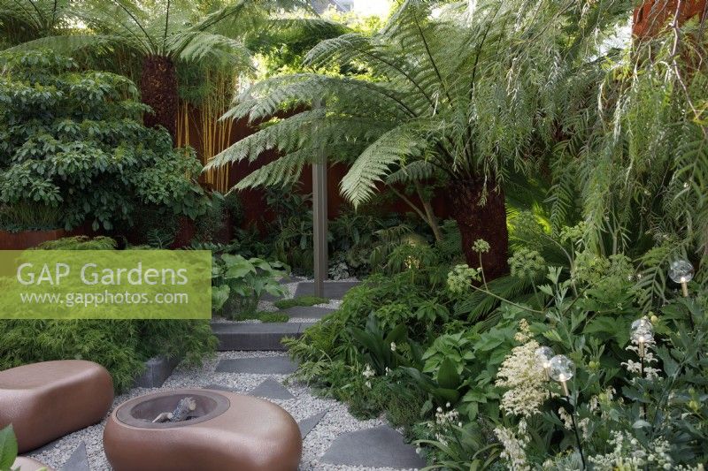 Lush and tropical planting in the Out of The Shadows garden which include Angelica archangelica, Dicksonia antarctica, bamboos with glass baubles lighting, the seating and fire bowl are curvaceous pebble shaped - Designer and Sponsor: Kate Gould Gardens