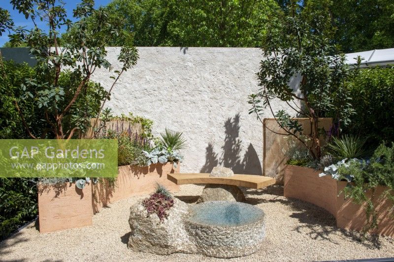 Mediterranean style garden with stone water feature and hand rendered stone effect wall.  Curved terracotta planters with  Arbutus, Salvia, Rosmarinus officinalis 'Prostratus' and Senecio candidans 'Angels Wings' -  A Mediterranean Reflection, RHS Chelsea Flower Show 2022 - Silver Medal