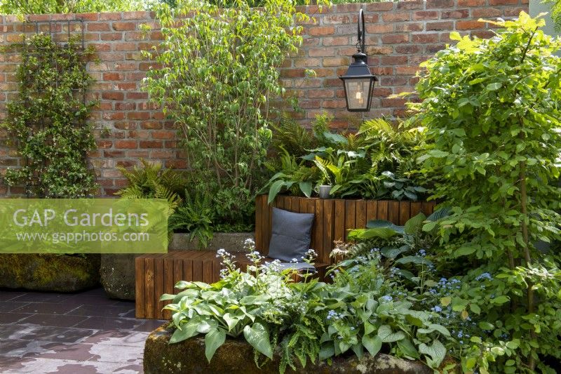 Stone troughs with shade loving plants including ferns, hosta and brunnera, Trachelospermum jasminoides trained against a brick wall with hanging lantern, wooden bench with cushion - The Enchanted Rain Garden, RHS Chelsea Flower Show 2022 - Silver Gilt Medal
