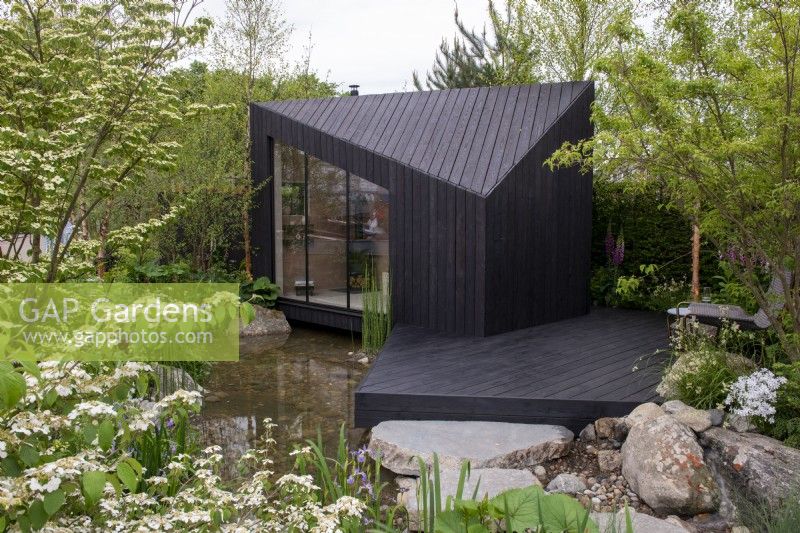 A charred larch cabin with decking overlooks a pond surrounded by Betula nigra -  A Garden Sanctuary by Hamptons,  RHS Chelsea Flower Show 2022