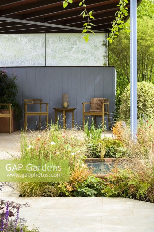 Garden with covered seating area with blue walls,  beside  pond planted with Iris 'White Swirl', Carex buchananii  with  green and  bronze planting  - SSAFA Sanctuary Garden. Designer: Amanda Waring