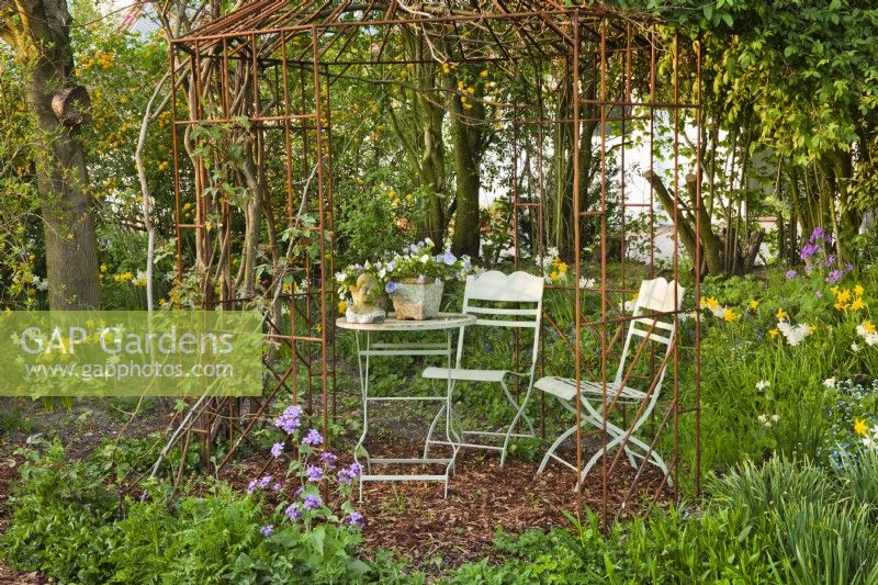 Metal garden furniture under arbour in spring garden with head statue and pots on the table.