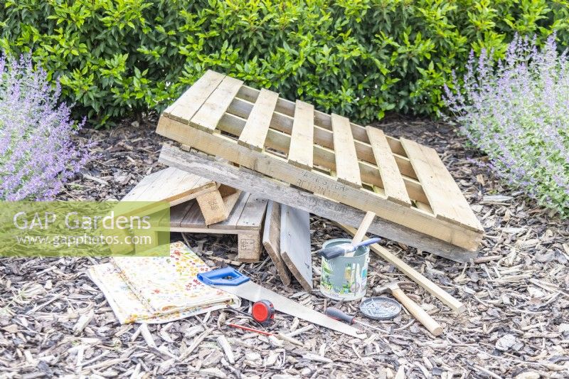 Pallets, planks, paint, paint brush, wax cloth, saw, pencil, tape measure, screwdriver, screws, hammer, nails and a thin wooden beam laid out on the ground