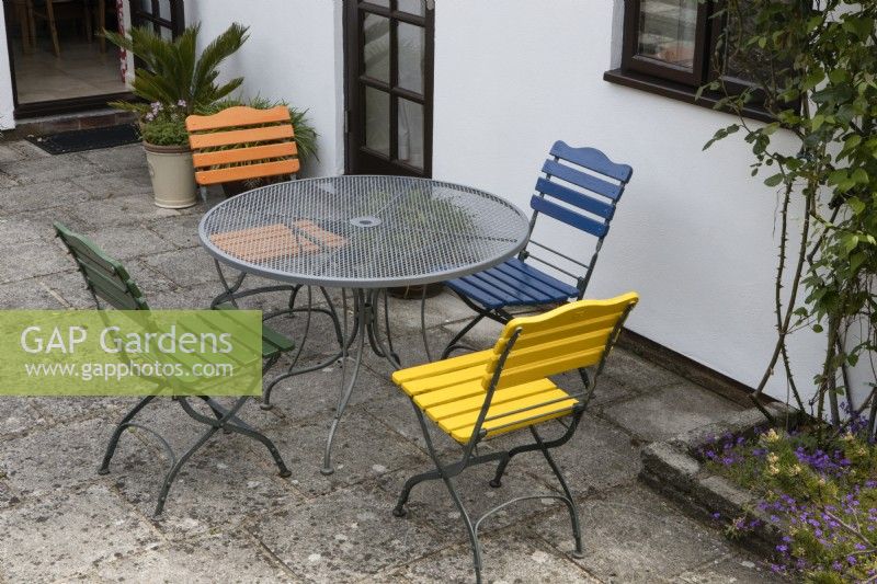 A round table with four different coloured seats on a patio area. Briar Cottage Garden. Devon NGS garden. Spring