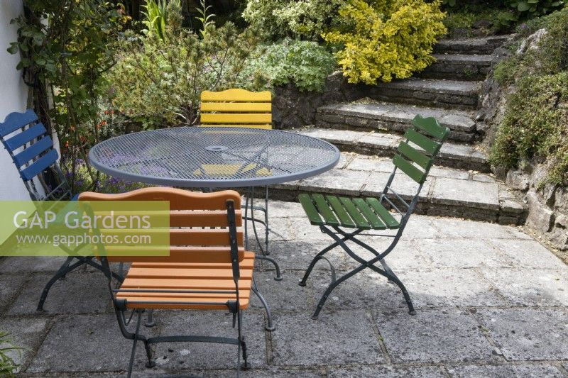 A round glass table sits on a patio area with four different coloured seats. Briar Cottage Garden. Devon NGS garden. Spring