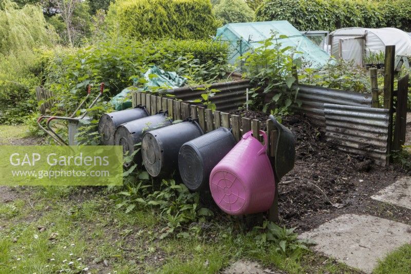 Five plastic trugs of various sizes hang beside a large compost bin with a wheelbarrow and various garden structures in the background. Lewis Cottage, NGS Devon garden. Spring.