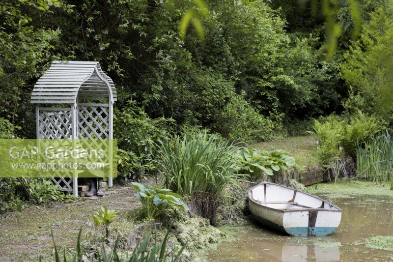 A wooden rowing boat on a pond with a wooden arbour seat on the edge and a pair of waders beside the seat. Lewis Cottage, NGS Devon garden. Spring.