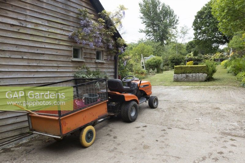 A Husqvarna lawn tractor and trailer beside a garden. Lewis Cottage, NGS Devon garden. Spring.