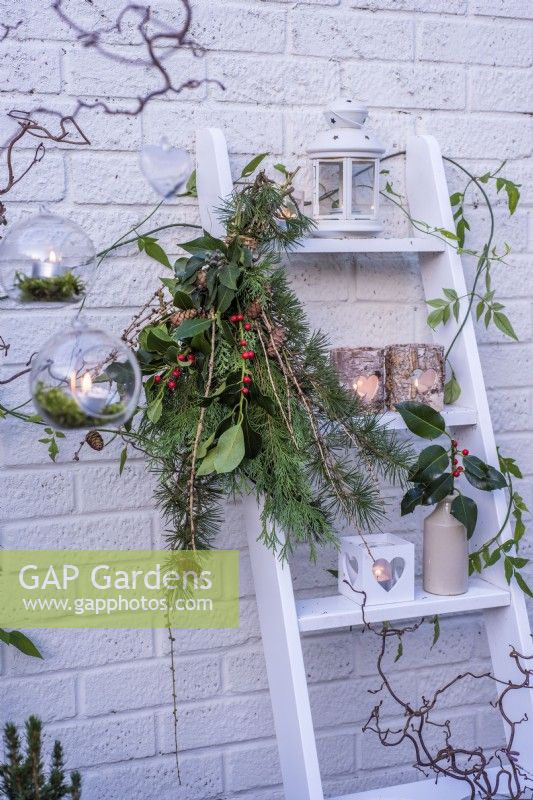 Tied bunch of evergreen foliage - conifers, ilex and Larix decidua cones attached to white ladder decorated with tealights