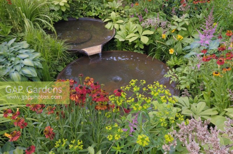 Water flowing between two staggered Torc Pots corten steel water bowls surrounded with colourful plants including helenium, euphorbia and astilbe.  The Daily Mail and RHS Planet-Friendly Garden, RHS Hampton Court Palace Garden Festival 2022.  Designer: Mark Gregory 