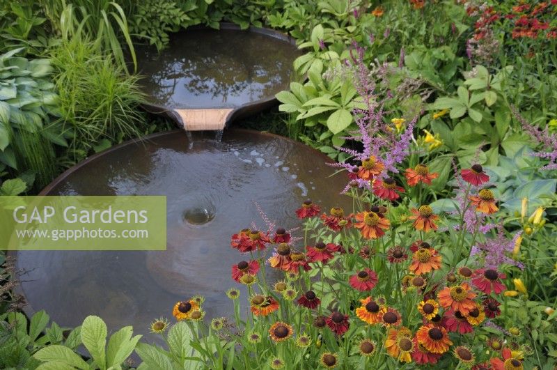 Water flowing between two staggered Torc Pots corten steel water bowls surrounded with colourful plants including helenium and astilbe.  The Daily Mail and RHS Planet-Friendly Garden, RHS Hampton Court Palace Garden Festival 2022.  Designer: Mark Gregory 
