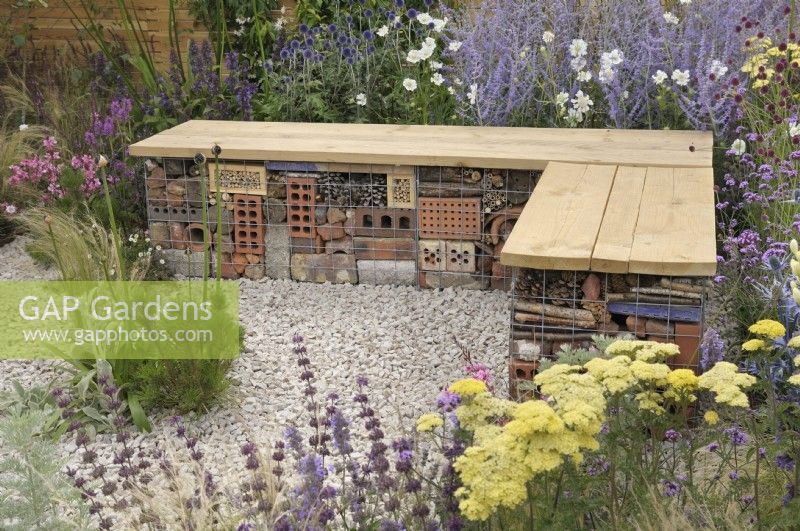 Bug or insect hotel packed inside a gabion basket with wooden bench top - Turfed Out Garden, RHS Hampton Court Palace Garden Festival 2022.  July.  Designer: Hamzah-Adam Desai  