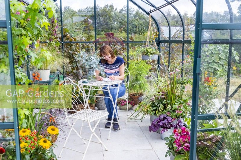 Woman sitting at table writing in a greenhouse filled with various plants and containers