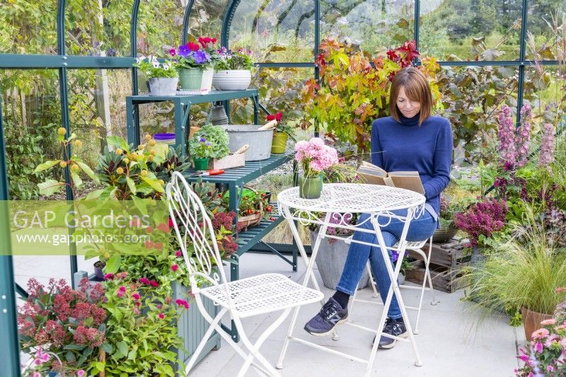 Woman sitting at a table reading in a greenhouse that has been filled with various plants and mixed containers