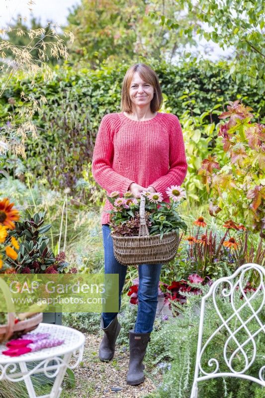 Woman carrying a basket containing Coprosma 'Inferno' and Echinacea 'Sunseekers Salmon' through garden