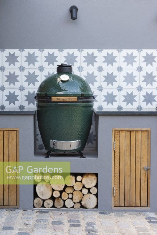 Detail of outdoor modern kitchen with Big green egg, grey walls with decorative wall tiles and storage with wood piles. 
