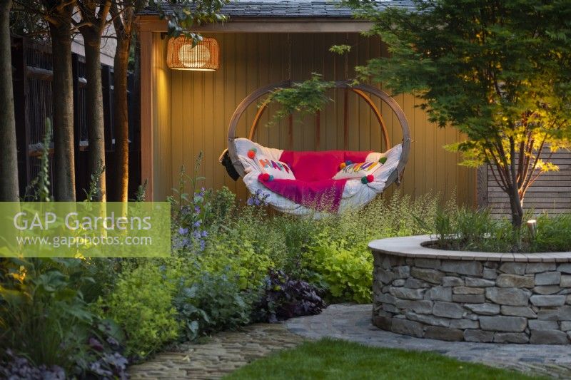 Illuminated outdoor reading room with hanging wooden bench next to the borders with purple and lime Heuchera. 
