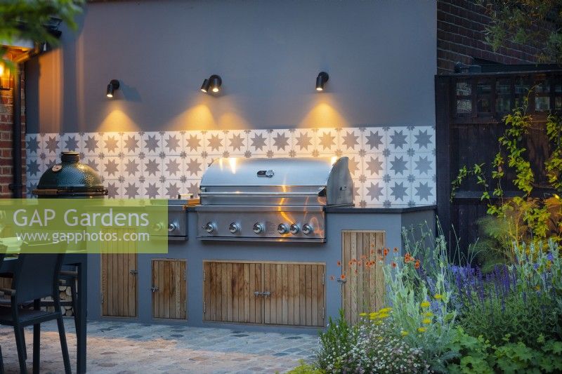 Illuminated outdoor kitchen with ornamental tiles, grey wall, bbq and storage.  