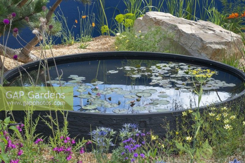 A circular raised pool is set into a gravel garden planted with colourful perennials amongst boulders.