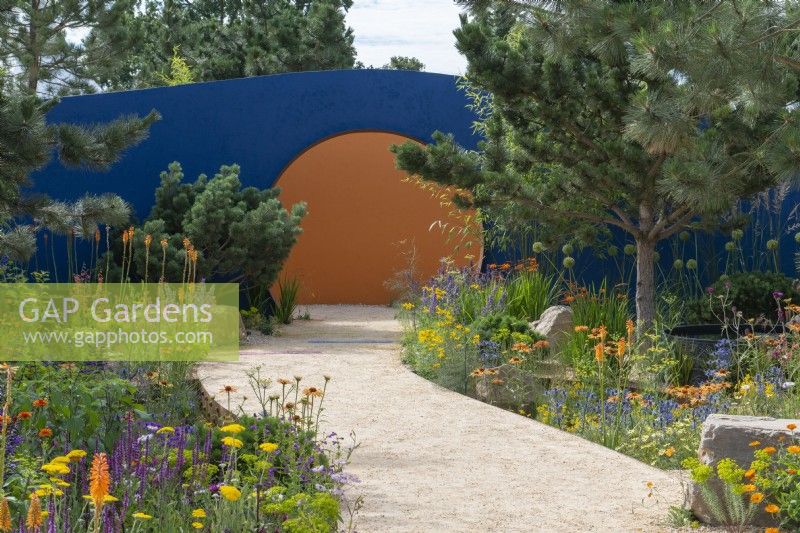 A broad path curves towards a circular opening in a blue-painted wall, passing between borders of pines and colourful perennials.
