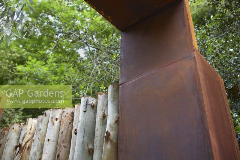 Corten Steel garden structure with Eucalyptus log fence. Summer.