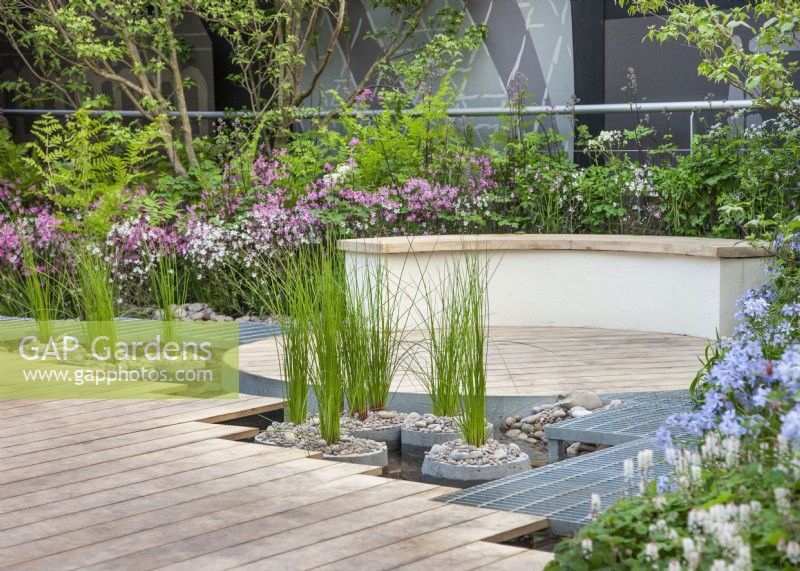 Caption: Modern garden design, decked surface near water with curved bench. Summer July