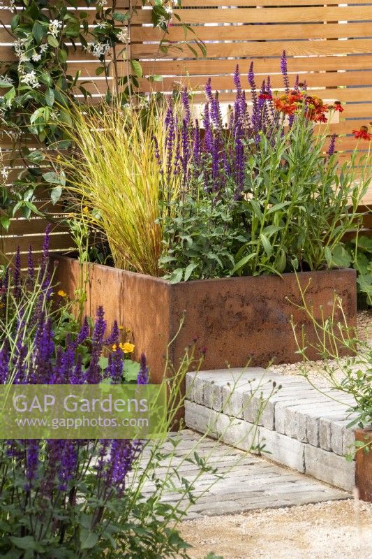 Rusted steel planters filled with perennials with benches, wooden fence, steps, and clay paver and gravel paths - RHS Hampton Court Palace Garden Festival, London, July 2022 - 'Lunch Break Garden', Best in Show Get Started Gardens - Designer Inspired Earth Design