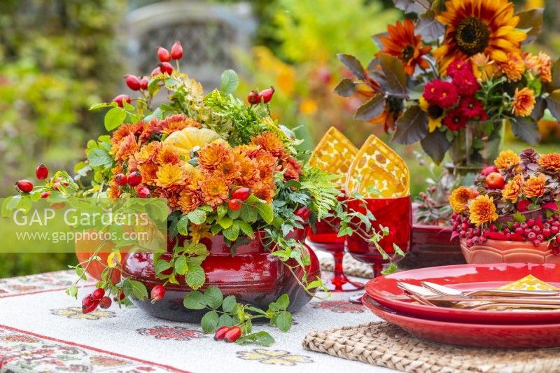 Red container with orange Chrysanthemums, Rosehips, Ivy, Ferns and small squash on a table in the garden
