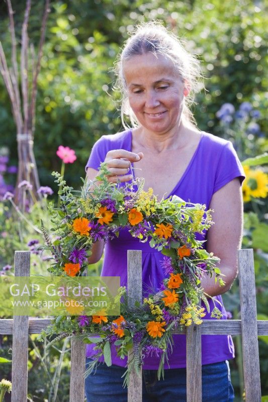 Woman hanging wreath made of edible flowers and herbs on fence to dry.