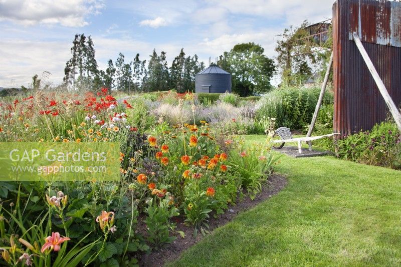 View of the borders at the Cottage Herbery with the vibrant red bed in the foreground, which planting includes Crocosmia 'Lucifer', Geum 'Totally Tangerine', Echinacea 'Cantaloupe' and Rudbeckia 'Cherokee Sunset' and further back we can see the derelict dutch barn and an old silo painted in black.