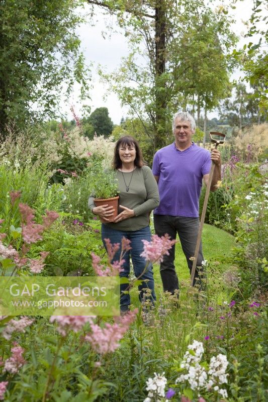 Couple standing by flower beds holding pot of Agastache 'Fleur' and a hoe