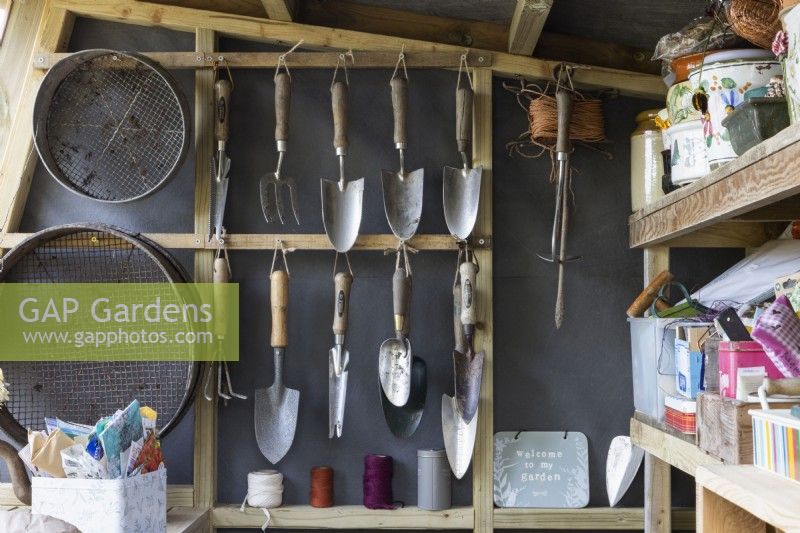 A neat garden shed with trowels and garden sieves hanging at one end and various gardening paraphernalia on shelves on the right hand side. Autumn.