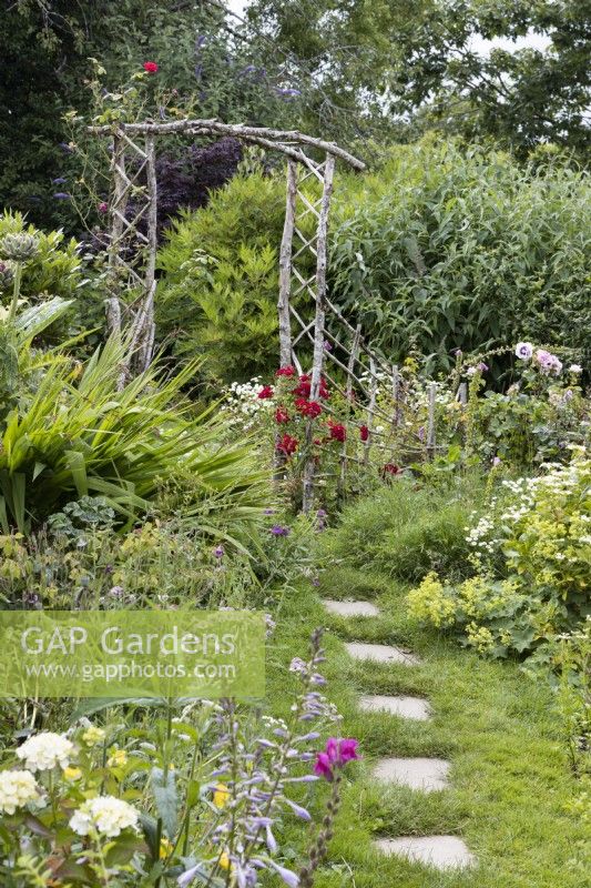 A paving slab path leads to a rustic, home made wooden arch with perennial planting either side and a rambling red rose, Rambling Rosie,  growing beside the arch.  Derryn Bank. June.