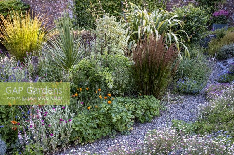 Winding gravel path leads through a mixed cottage style garden border, with grasses, annuals and perennials mixed together. Large Phormiums at the back give height.