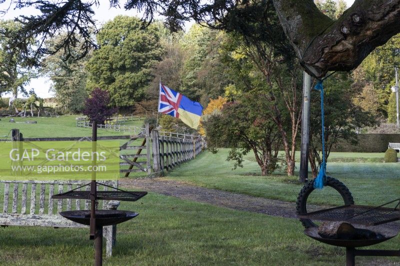 Several barbecues with an old tyre rope swing and wooden bench in foreground with a wooden post and rail gate and fence in background with a Union jack and Ukraine flag flying. Regency House, Devon NGS garden. Autumn