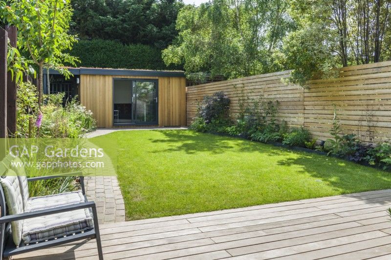 Garden office at the end of a contemporary garden with lawn, stone block path and slatted timber fencing. June