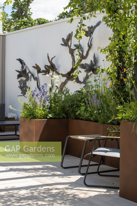 Raised Corten steel planters surround a decked seating area with wall sculpture by Emma Rodgers - John King Brain Tumour Foundation Garden, RHS Hampton Court Palace Garden Festival 2022