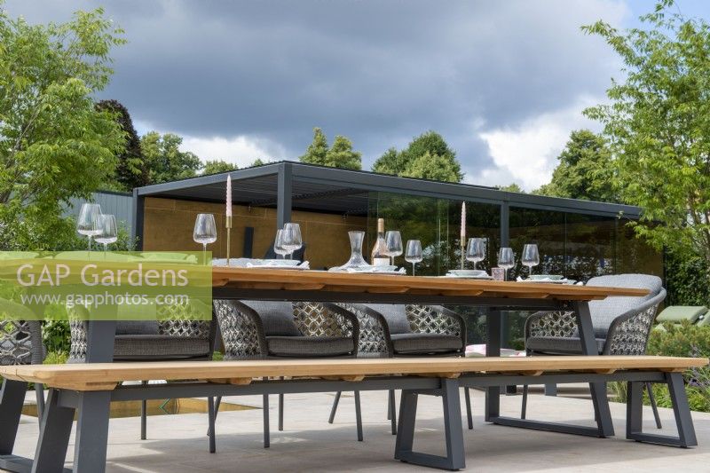 Outdoor dining table with benches and metal pergola behind - The Outdoor Living Garden, RHS Hampton Court Palace Garden Festival 2022