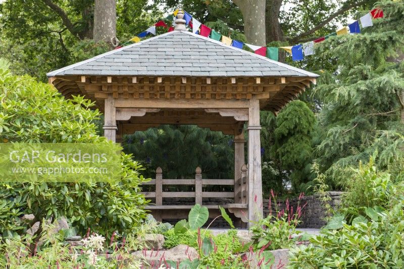 Wooden shelter resembling Nepalese religious building, with prayer flags, planting includes large Rhododendrons and Persicaria amplexicaulis - The Trailfinders 50th Anniversary Garden, RHS Chelsea Flower Show 2021