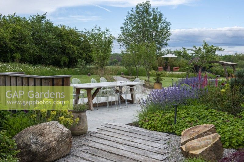 Large boulders rest beside timber planks laid diagonally in stone chippings, leading to a contemporary terrace edged in hardy geranium, catmint and lupin.
