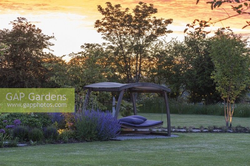 Seen at sunset, a swingseat sits on stone chippings between a lawn and a border planted with perennials.