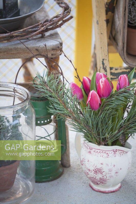 Close up and detail of a modern balcony with yellow railings in January.  Chamaecyparis pisifera 'Boulevard' in a glass jar next to a lantern and a jug with pine branches and tulips. 