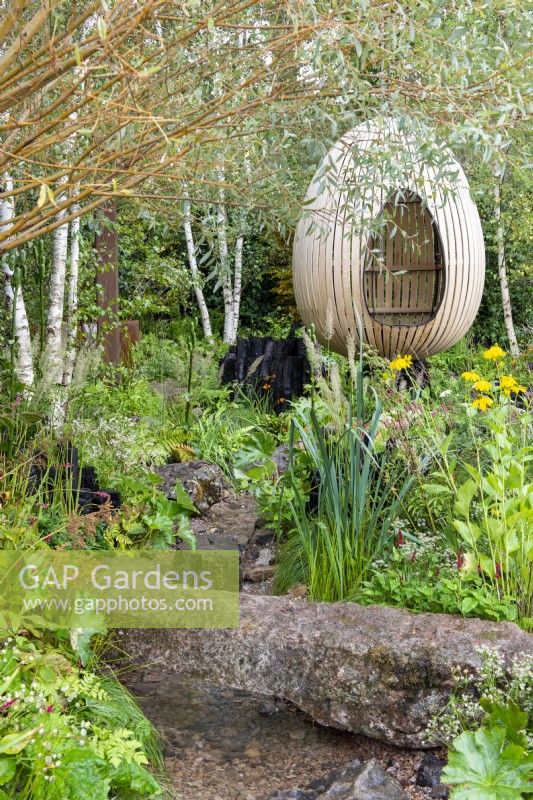 Bent oak swinging hide, suspended over naturalistic style planting with stream and stone bridge - The Yeo Valley Organic Garden, RHS Chelsea Flower Show 2021