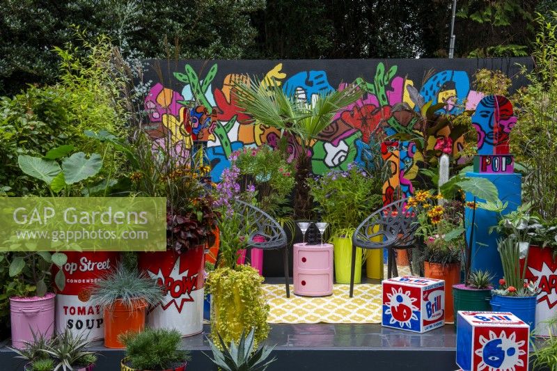 Planting in colourful containers surround seating area with black chairs and wall mural behind - Pop Street Garden, RHS Chelsea Flower Show 2021